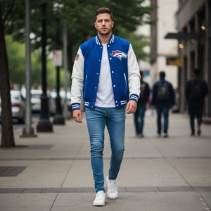 Man wearing a blue and white varsity jacket with a logo, white shirt, blue jeans, and white sneakers on a city street.