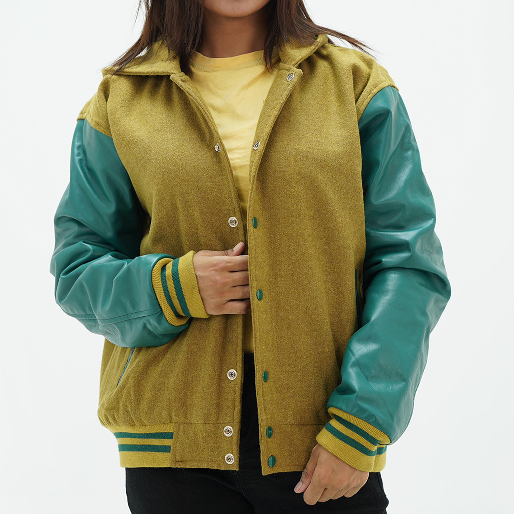 A woman wearing a camel body and green leather sleeves varsity jacket with a hood. The jacket features a yellow collar, green stripes on the sleeves, and coated snap-button fastening.
