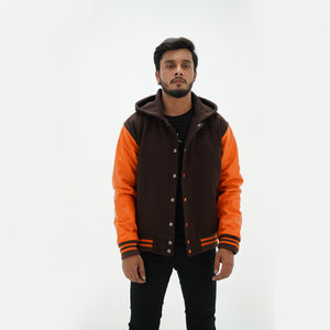 A male model wearing a chocolate brown varsity jacket with old gold leather arms and a hoodie. The jacket has orange ribbing and old gold stripes, with a front snap fastening and a collar lined with old gold quilting.