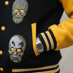 Black and yellow varsity jacket with skull designs and a watch on a neutral background