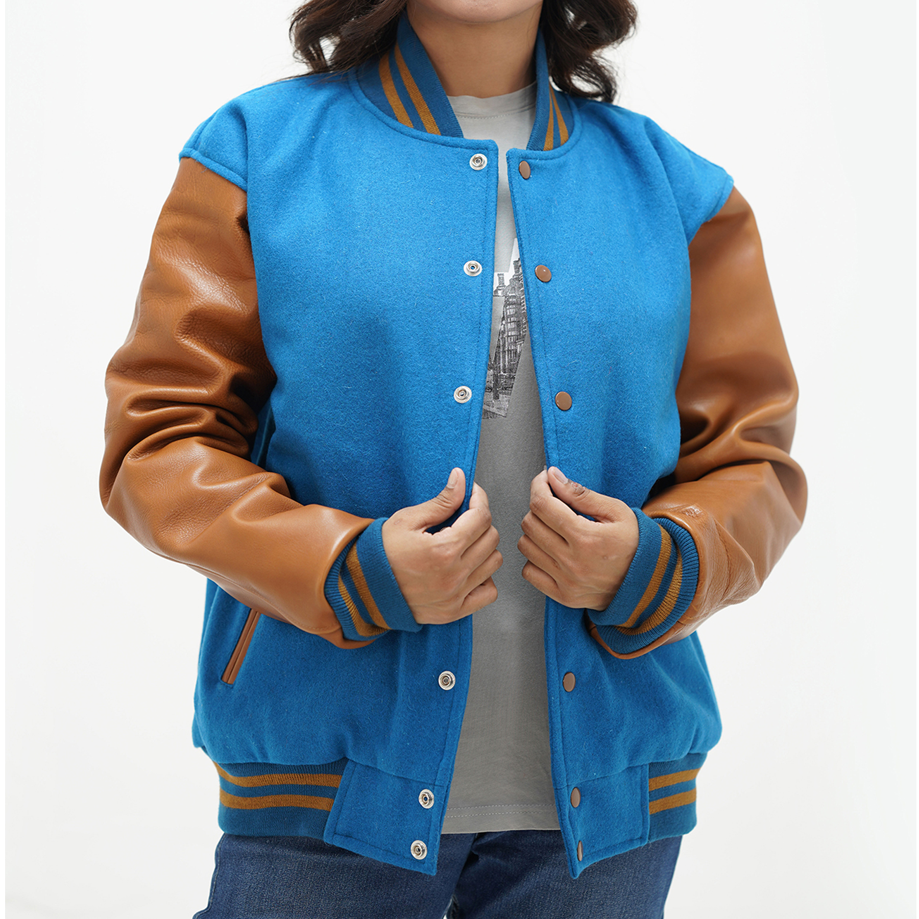 A woman wearing a sky blue wool letterman jacket with brown leather sleeves and a brown collar, featuring ribbed cuffs and a front with coated snaps. She is also wearing a grey shirt and blue jeans.