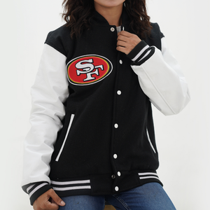 A woman wearing a black and white 49ers letterman jacket with leather sleeves and a hoodie. The jacket features a black body with white stripes and a white collar.