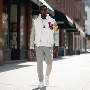 Man wearing a white varsity jacket with red lettering and gray pants walking on a city street.